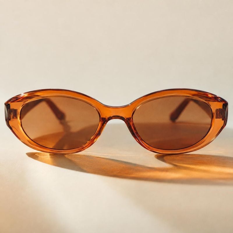 sunglasses packshot product photo: Amber-toned oval sunglasses rest on a warm beige surface, with crisp directional sunlight casting a long shadow and creating…