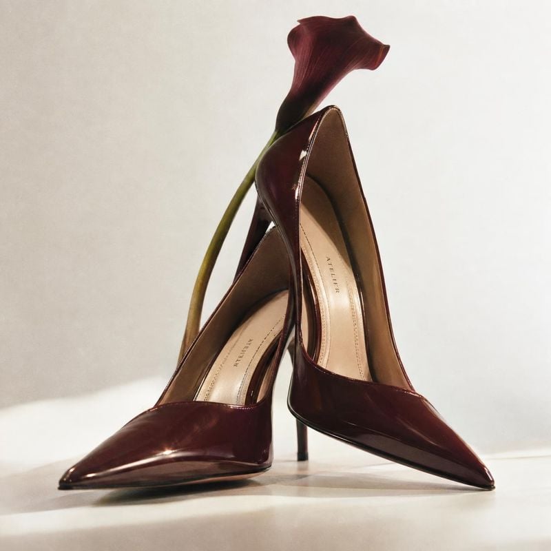 burgundy patent pumps fashion product photo: Glossy burgundy pointed-toe pumps are artfully arranged with a deep red calla lily against a softly lit neutral st…