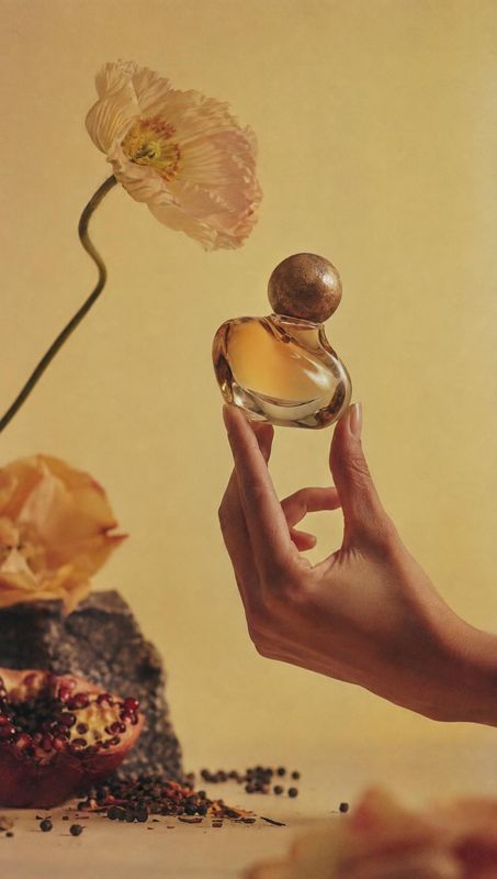 perfume bottle product in hand product photo: A hand presents an amber perfume bottle among dried flowers and scattered botanicals against a warm yellow studio…