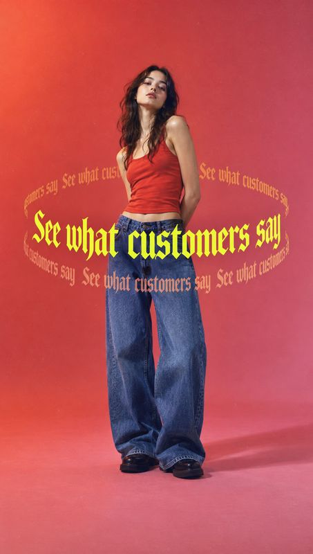 wide-leg jeans look book product photo: A model poses in a red tank top, loose blue jeans and dark shoes against a warm red-pink studio backdrop, with bold yel…