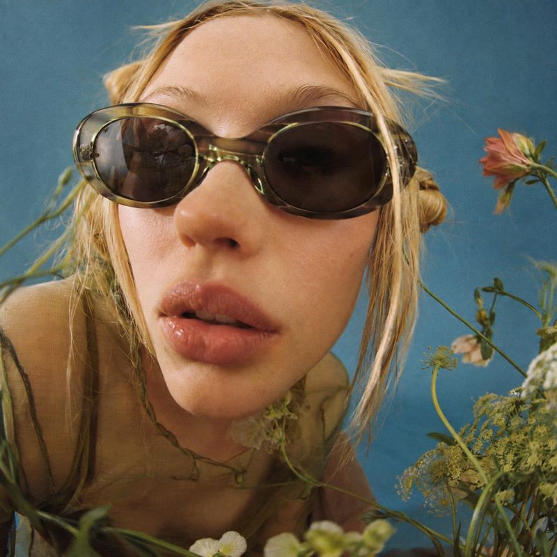 sunglasses model try-on product photo: A close, softly sunlit fashion portrait shows a blonde woman wearing oversized dark sunglasses amid delicate wildflowers…