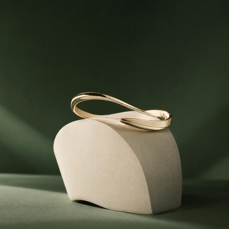 gold bracelet close-up detail product photo: A polished gold bracelet rests on a sculptural cream display against a deep green studio backdrop, with warm direc…