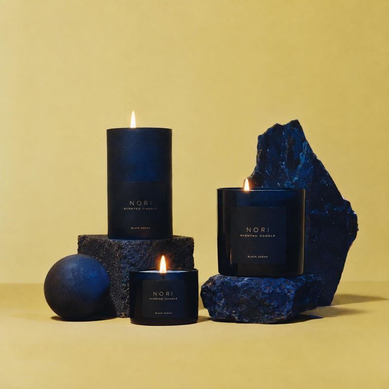scented candles black friday product photo: Three lit black candles are arranged among dark blue rocks and a sphere against a warm yellow studio backdrop, crea…