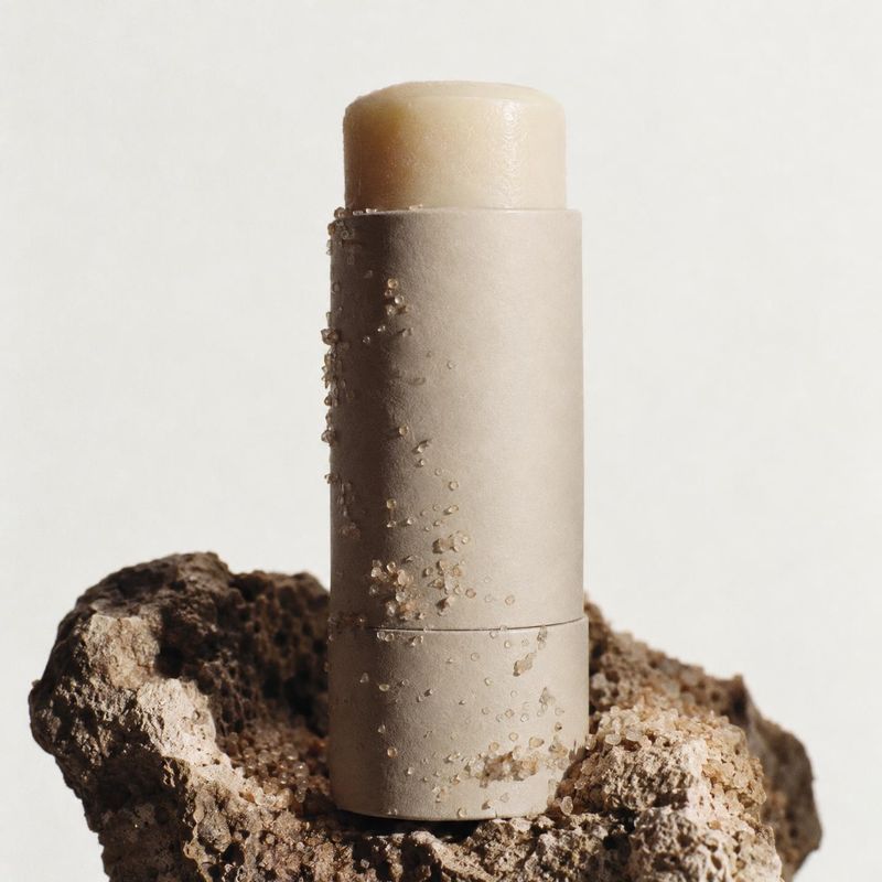 skincare balm stick close-up detail product photo: A minimalist beige balm stick stands upright on a rugged mineral rock, with scattered crystals and soft stud…