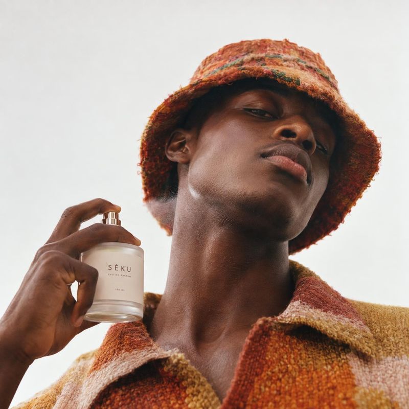 perfume model try-on product photo: A low-angle portrait shows a man in a textured rust-colored hat and coat holding a perfume bottle near his neck against a s…