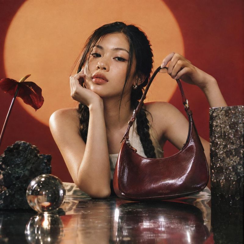 leather shoulder bag model try-on product photo: A model poses with a glossy burgundy shoulder bag against a warm orange-and-red studio backdrop, with sculptur…