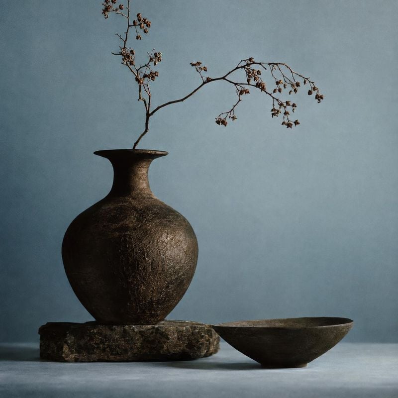 ceramic vase and bowl home product photo: A dark, textured ceramic vase with a delicate dried branch and a shallow bowl is arranged against a muted blue studio…