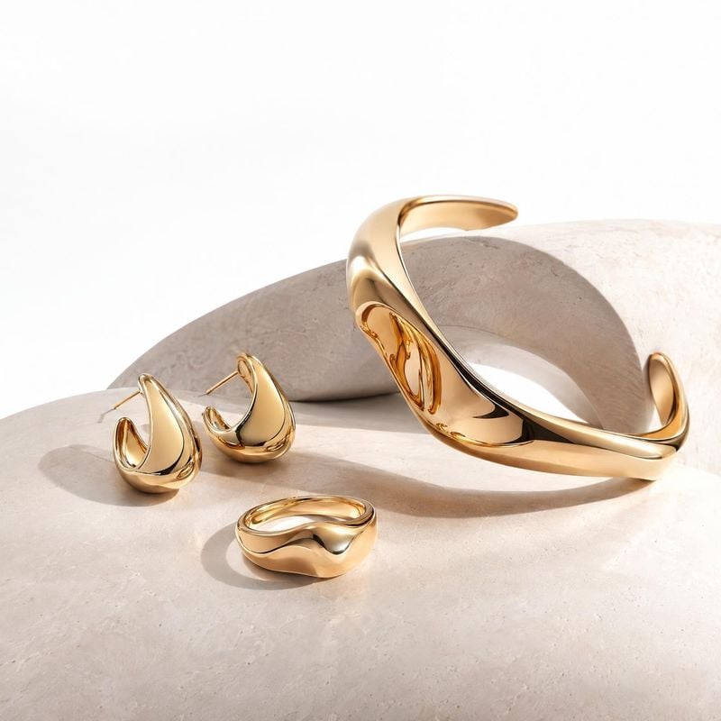 gold-tone jewelry set close-up detail product photo: A sculptural gold-tone cuff, earrings and ring are arranged on pale stone forms in bright, warm studio lig…