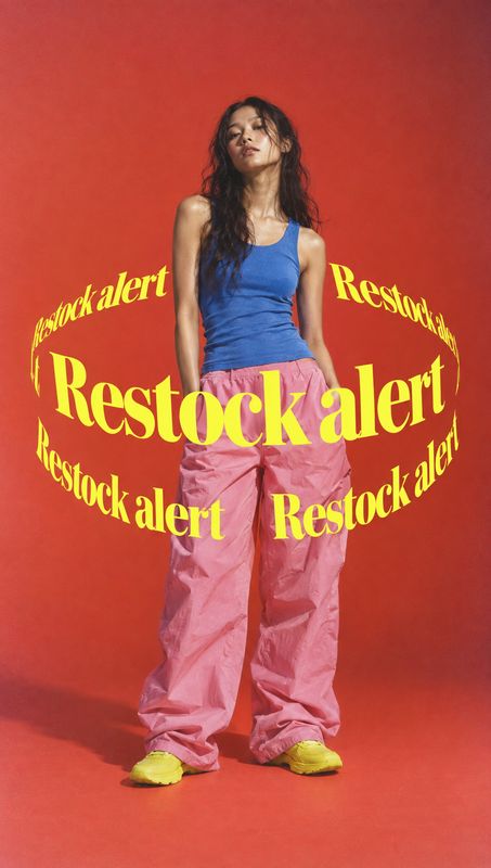 pink trousers look book product photo: A model poses in a blue tank top, oversized pink trousers and yellow shoes against a vivid red studio backdrop, encircle…