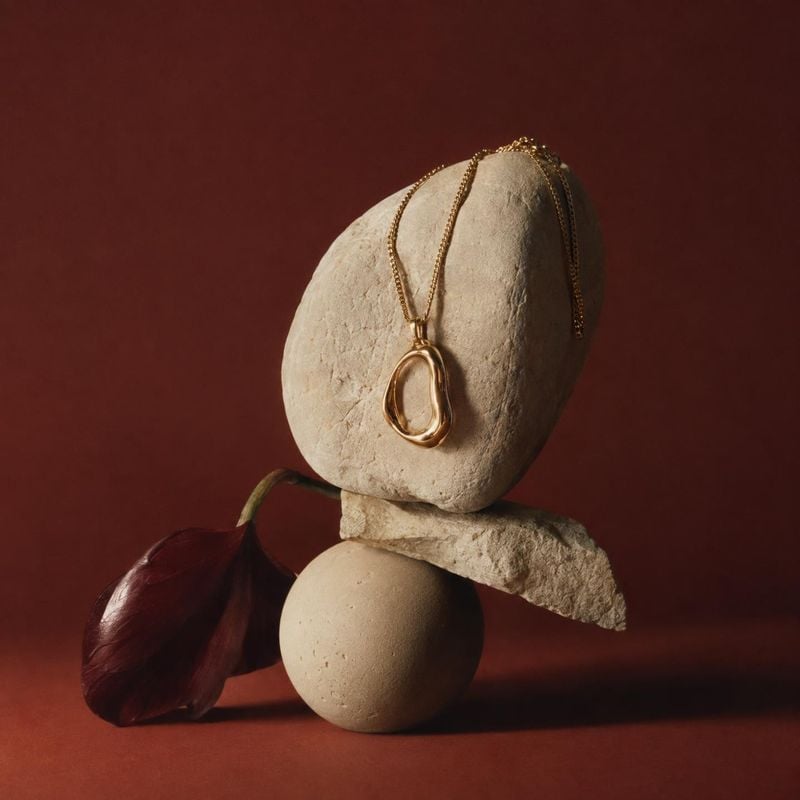 gold pendant necklace jewelry product photo: A sculptural gold pendant necklace drapes over pale stone forms beside a deep burgundy leaf, styled against a matc…