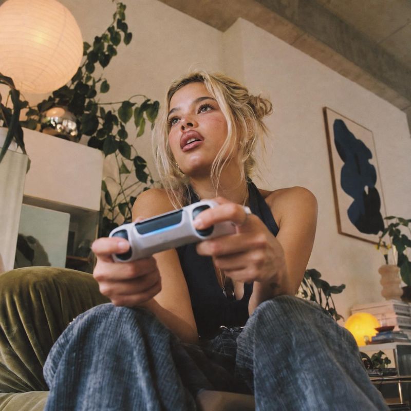 game controller lifestyle scene product photo: A blonde woman in a black top and loose jeans holds a game controller in a warmly lit, plant-filled living room,…