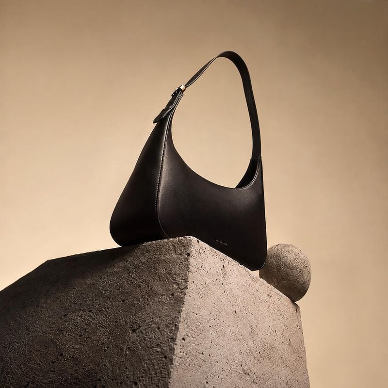 black leather shoulder bag jewelry product photo: A sculptural black shoulder bag rests on a rough stone form against a warm beige studio backdrop, lit with dr…