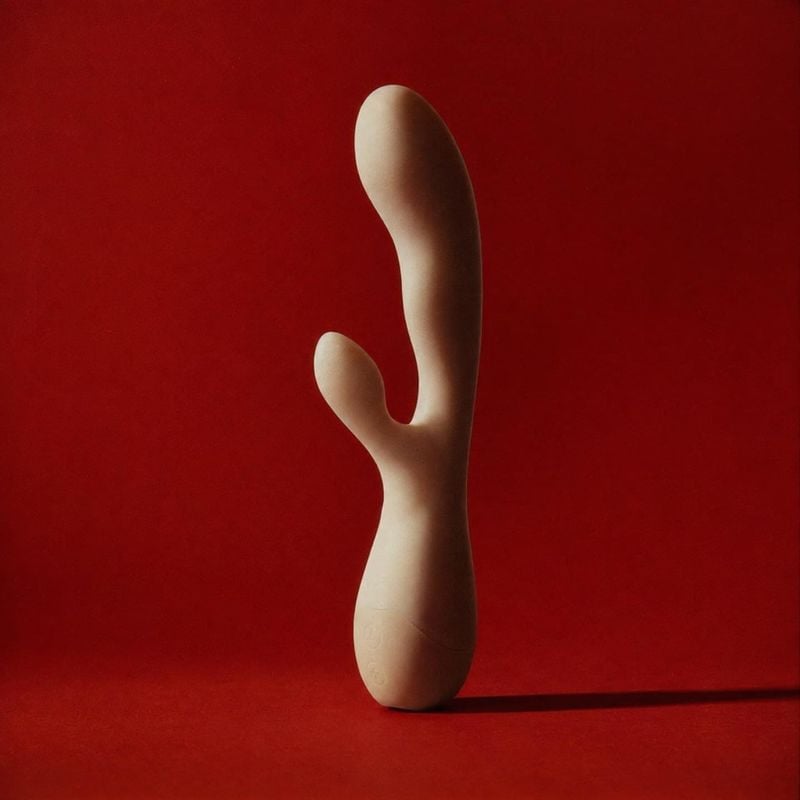 personal massager packshot product photo: A sculptural beige personal massager stands alone against a saturated red studio backdrop, lit dramatically with a lo…