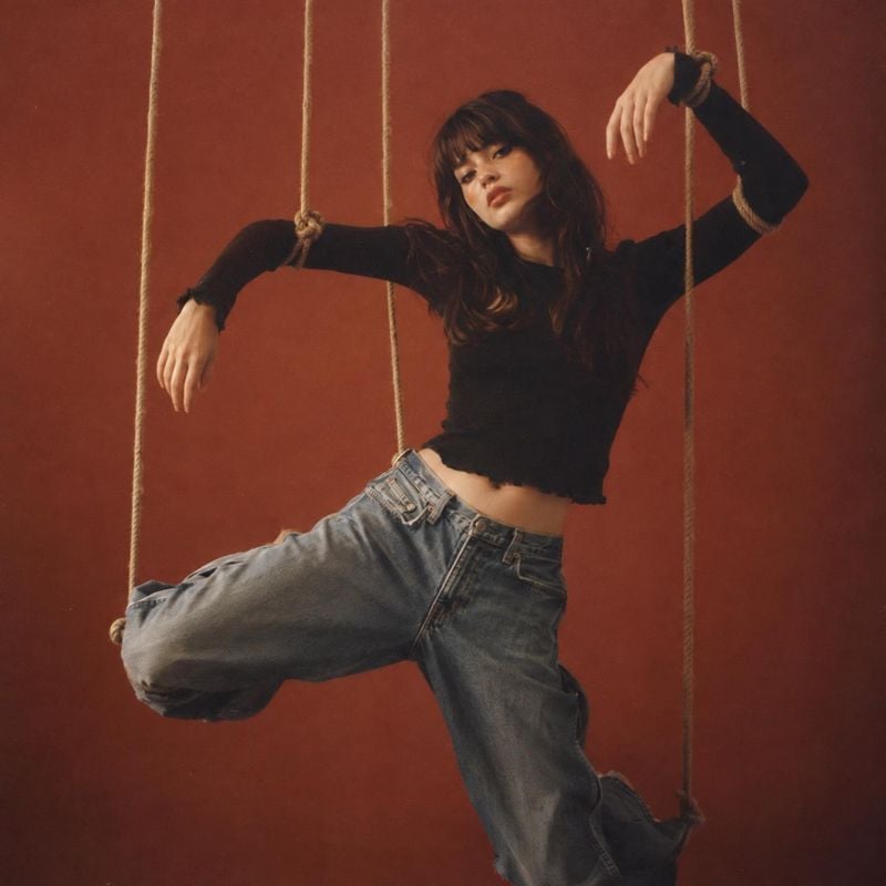 jeans and black top action & motion product photo: A model poses dynamically among hanging ropes in relaxed blue jeans and a fitted black top against a warm te…
