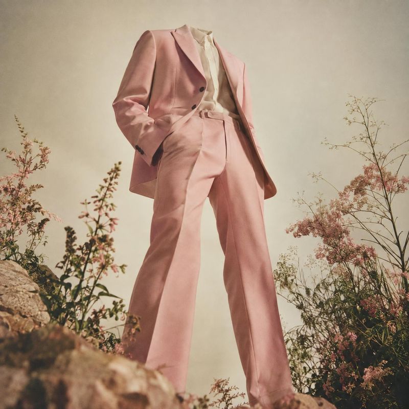 pink suit ghost mannequin product photo: A model wears a tailored blush-pink suit among delicate flowering plants, photographed from a low angle in soft, muted…