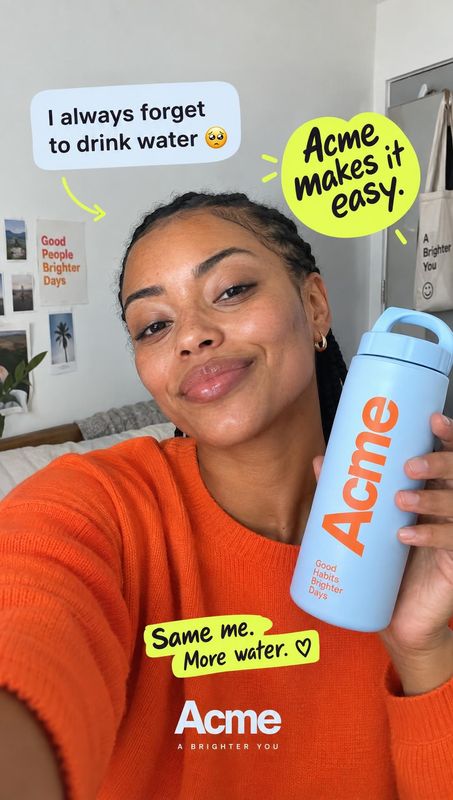 water bottle lifestyle scene product photo: A woman in an orange sweater smiles at the camera while holding a light-blue water bottle in a bright bedroom, with…