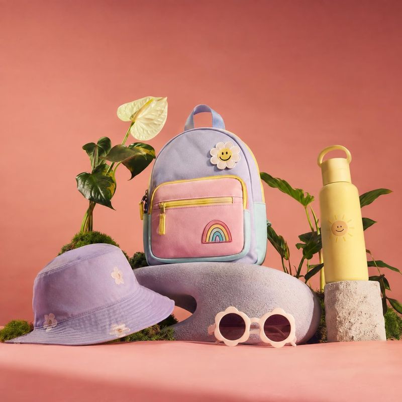 pastel children's backpack fashion product photo: A pastel backpack, bucket hat, sunglasses and water bottle are arranged among greenery on sculptural props ag…