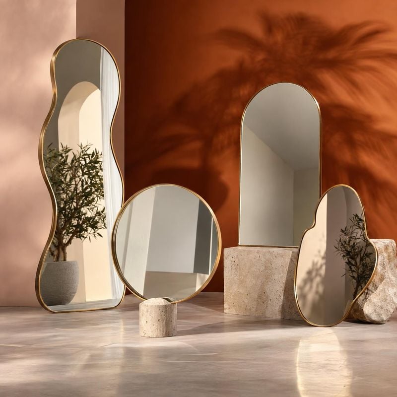 decorative mirrors group lineup product photo: A collection of gold-framed mirrors in organic shapes is arranged among stone blocks and a potted plant in a war…