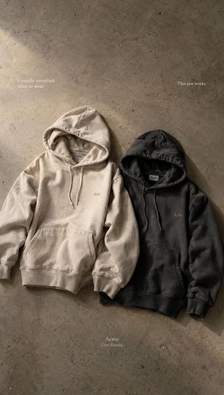 hoodies flat lay product photo: Two neutral-toned hoodies are arranged on a textured concrete floor, lit with soft directional light for a minimal, relaxed app…