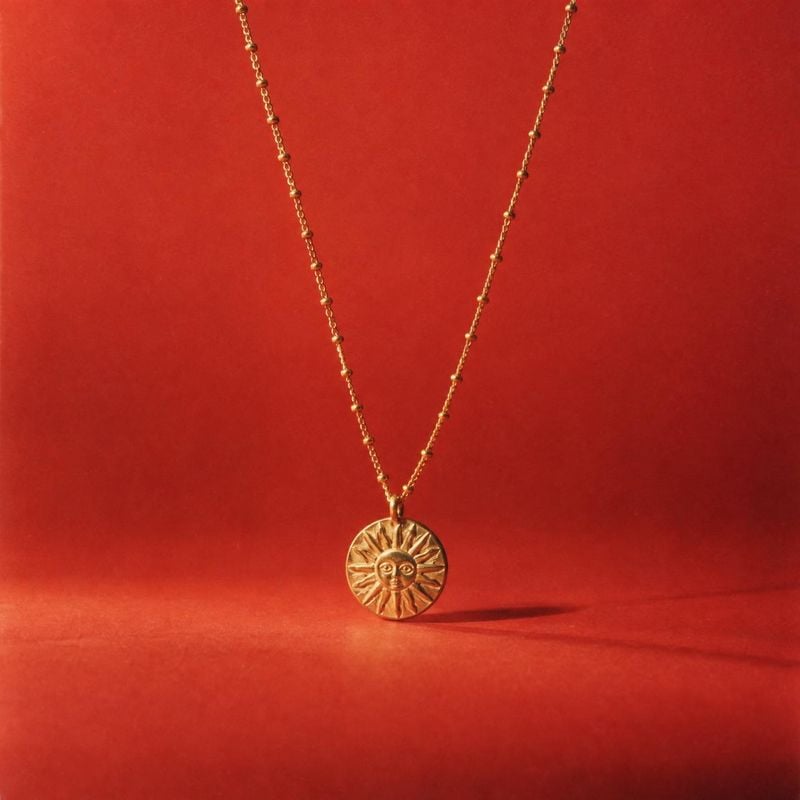 sun pendant necklace packshot product photo: A gold sun-face pendant necklace hangs against a rich red studio backdrop, with warm directional light creating a…