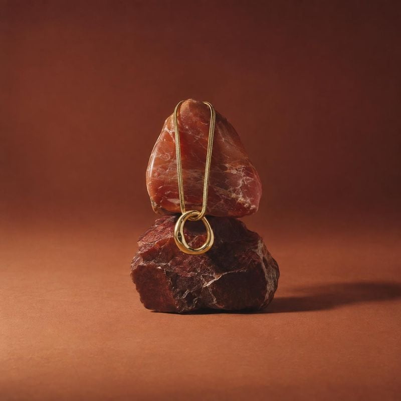 gold necklace close-up detail product photo: A delicate gold necklace is draped over rough reddish stones against a warm rust-colored studio backdrop, with sof…