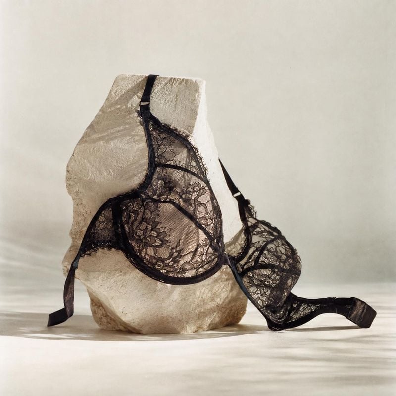 black lace bra close-up detail product photo: A black lace bra is draped over a pale sculptural stone against a softly lit neutral backdrop, creating an intima…