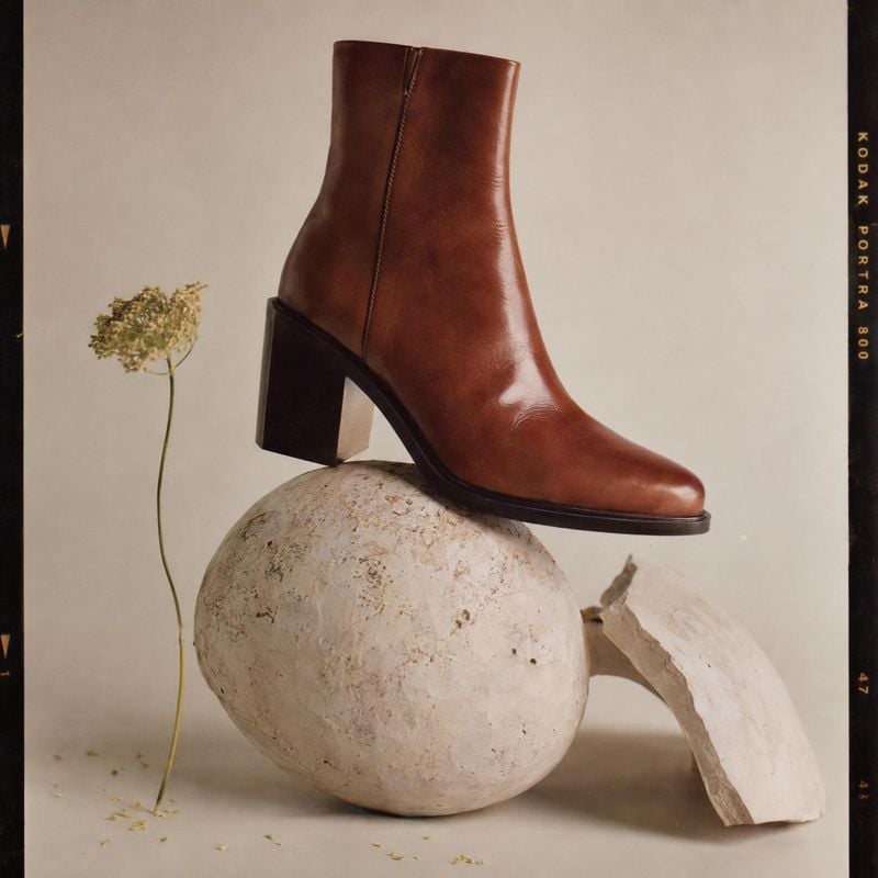 brown leather ankle boot fashion product photo: A polished brown leather ankle boot balances on sculptural pale stone forms beside a delicate dried stem, style…
