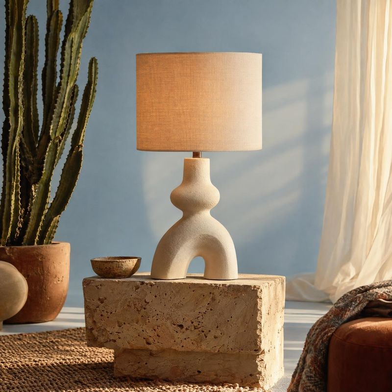 table lamp room set product photo: A sculptural cream table lamp sits on a stone plinth among a potted cactus and woven textures, lit by warm sunlight in a cal…