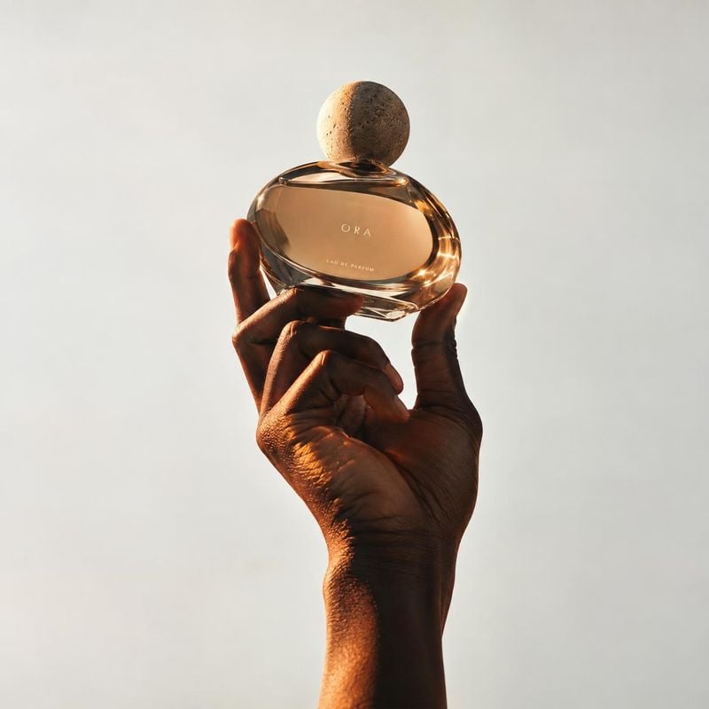 perfume product in hand product photo: A hand presents an amber perfume bottle against a pale seamless background, with warm directional light and a minimal, r…