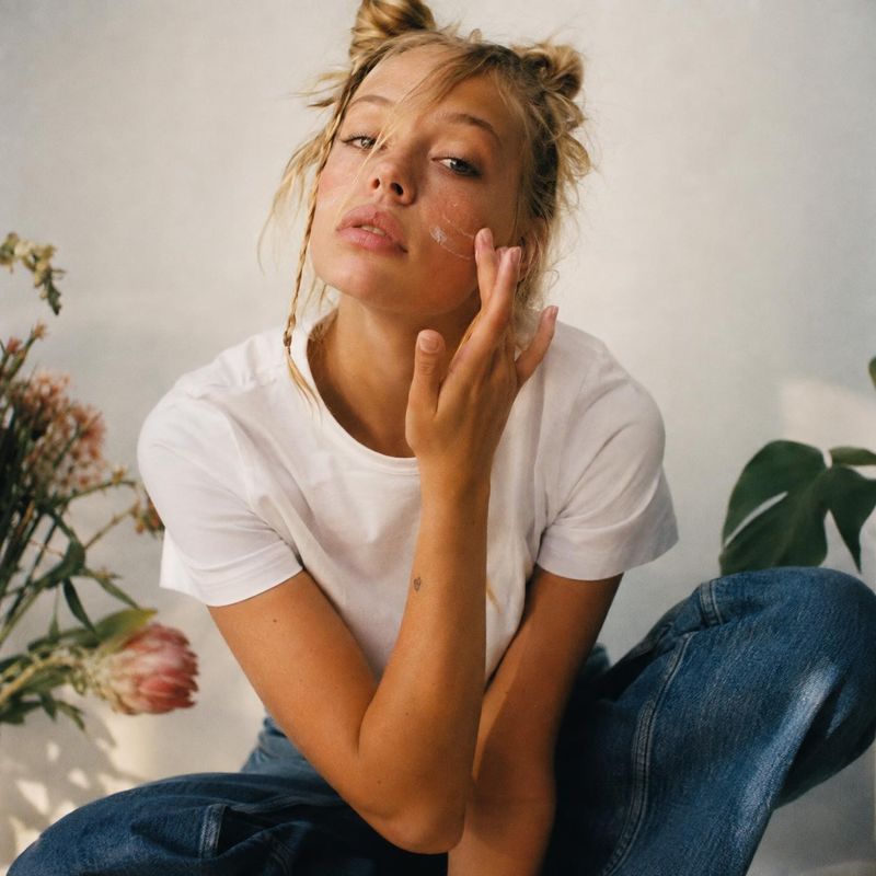 beauty close-up product photo: A woman in a white T-shirt and blue jeans gently applies cream to her cheek in a softly lit interior with leafy plants and flowe…
