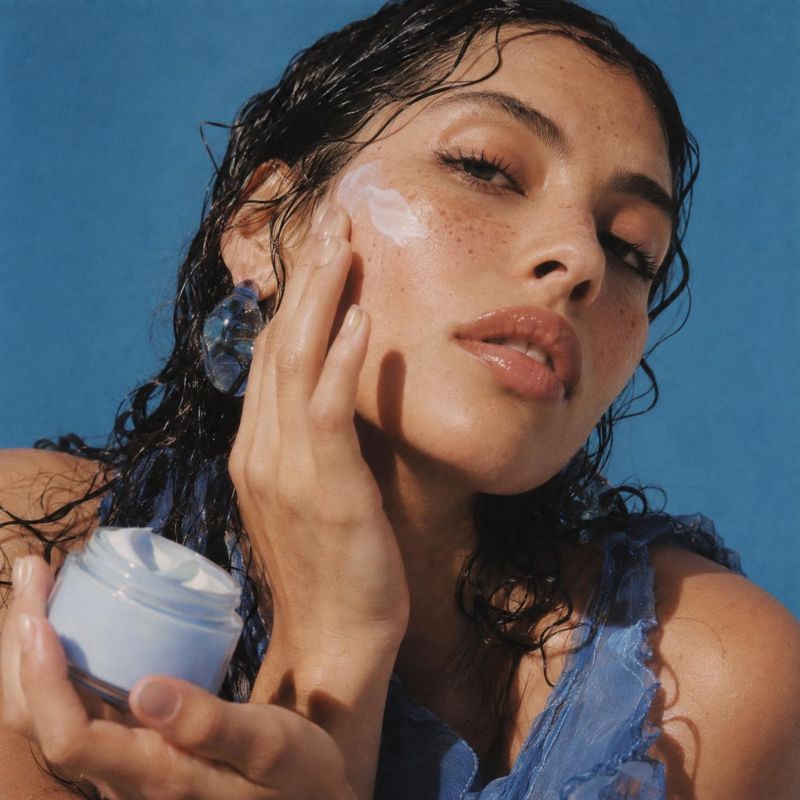face cream beauty close-up product photo: A close-up beauty portrait shows a woman applying cream to her cheek while holding an open jar against a vivid blue s…