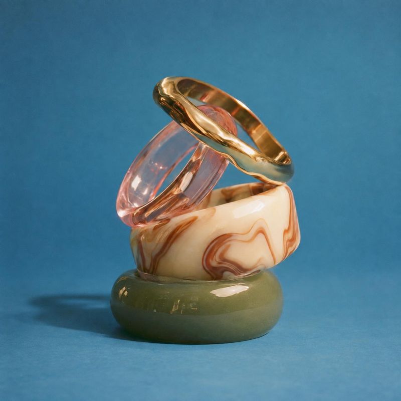 stacked rings close-up detail product photo: A sculptural stack of gold, pink, marbled cream, and olive rings is arranged against a rich blue studio backdrop,…