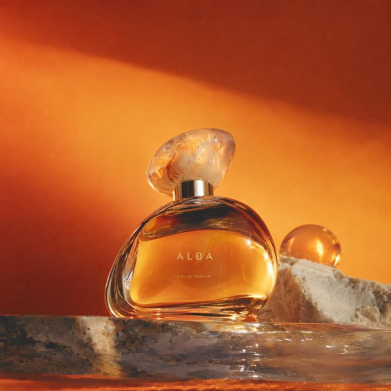 perfume close-up detail product photo: An amber perfume bottle rests on a textured stone surface beside a small orb, dramatically lit against a warm orange bac…