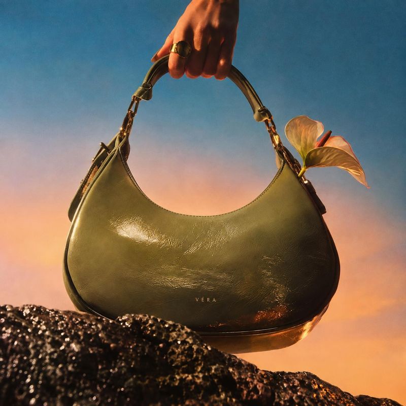 handbag product in hand product photo: A glossy olive-green handbag is held by its handle above dark rocks, with a flower tucked beside it against a softly glo…