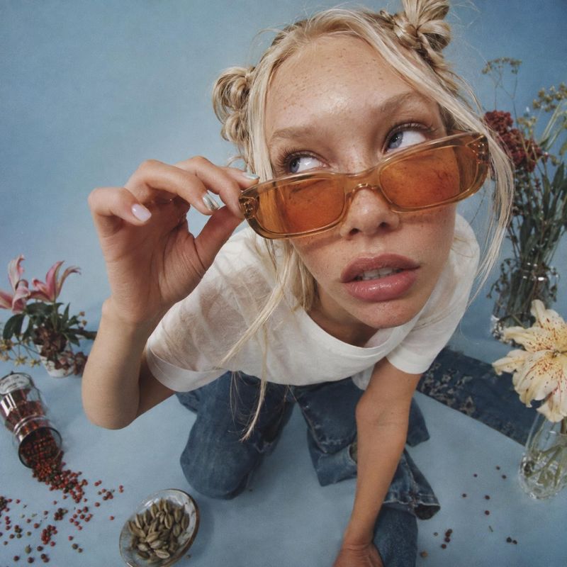 sunglasses model try-on product photo: A blonde model leans toward the camera in amber-tinted sunglasses, a white top and jeans, posed among scattered flowers…