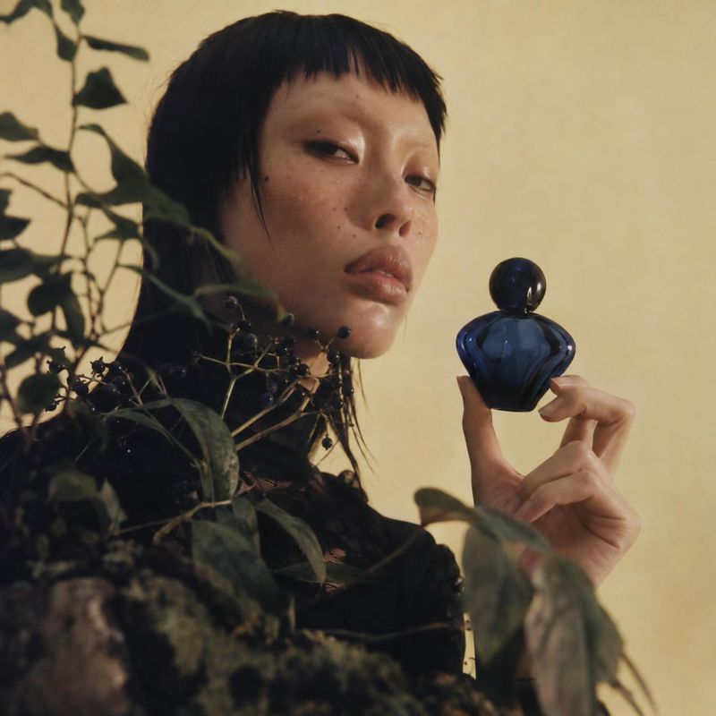 perfume bottle beauty close-up product photo: A model with short dark hair holds a deep-blue perfume bottle among shadowy botanical foliage against a warm beig…