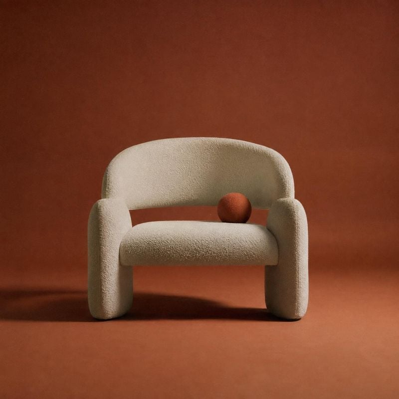 upholstered armchair packshot product photo: A sculptural cream bouclé armchair with a rust-colored sphere sits against a seamless terracotta backdrop, lit sof…