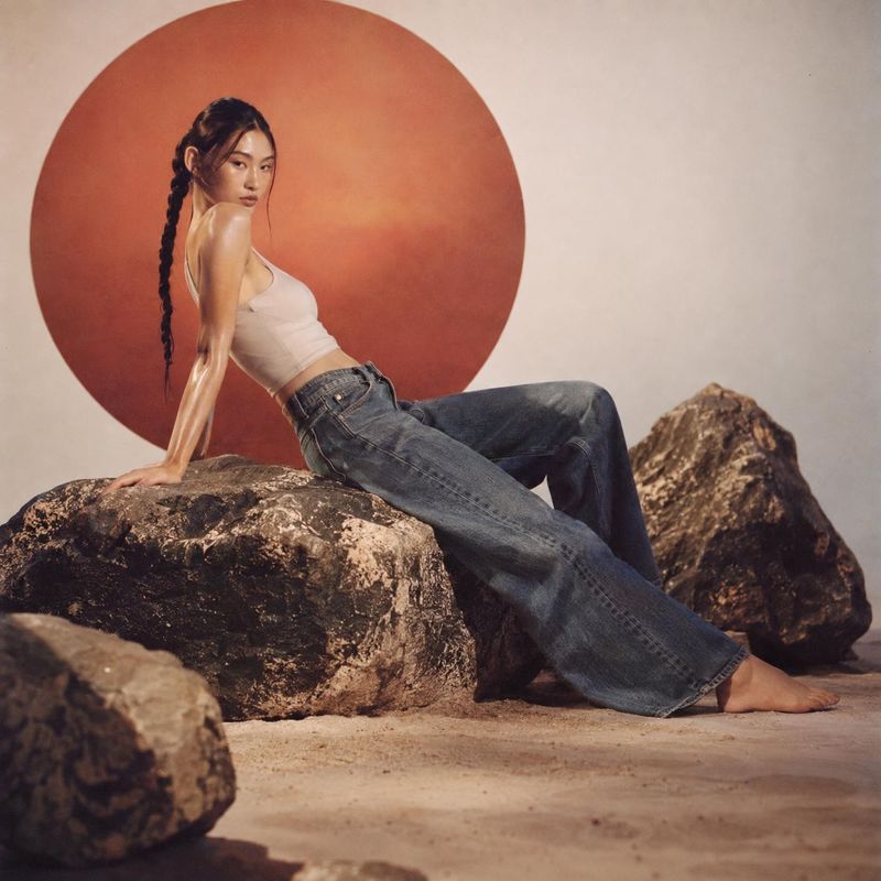 denim jeans model try-on product photo: A model in a white cropped tank and loose denim jeans reclines barefoot on sculptural rocks against a warm terracotta b…