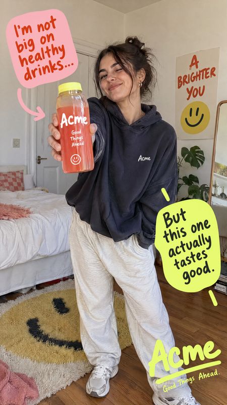 bottled drink food and drink product photo: A smiling woman in a dark hoodie and light trousers presents a pink bottled drink in a cozy bedroom, with bright ha…