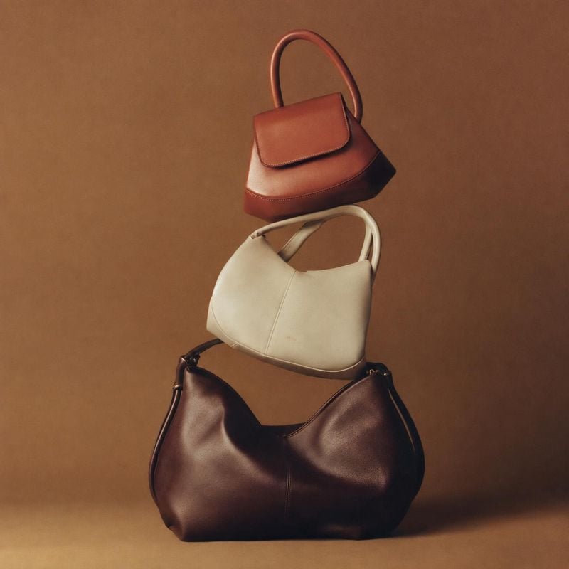 handbags group lineup product photo: Three sculptural leather handbags in warm brown, cream, and deep chocolate are stacked against a seamless terracotta-brown…