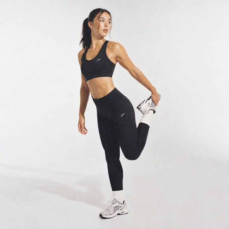black sports bra and leggings model try-on product photo: A woman in a black athletic set and white sneakers holds a standing quad stretch against a softly lit…