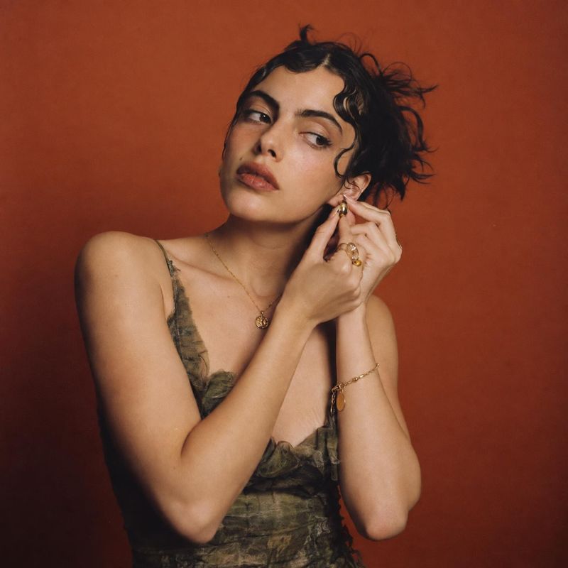 gold jewelry model try-on product photo: A woman in a textured olive dress adjusts a gold earring against a warm rust-red studio backdrop, with soft directiona…