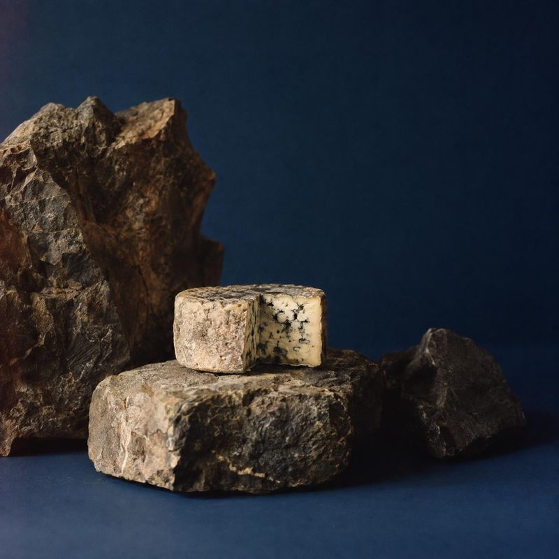 blue cheese food and drink product photo: A small wedge of blue cheese rests on rugged stone against a deep navy backdrop, with warm directional light and an e…