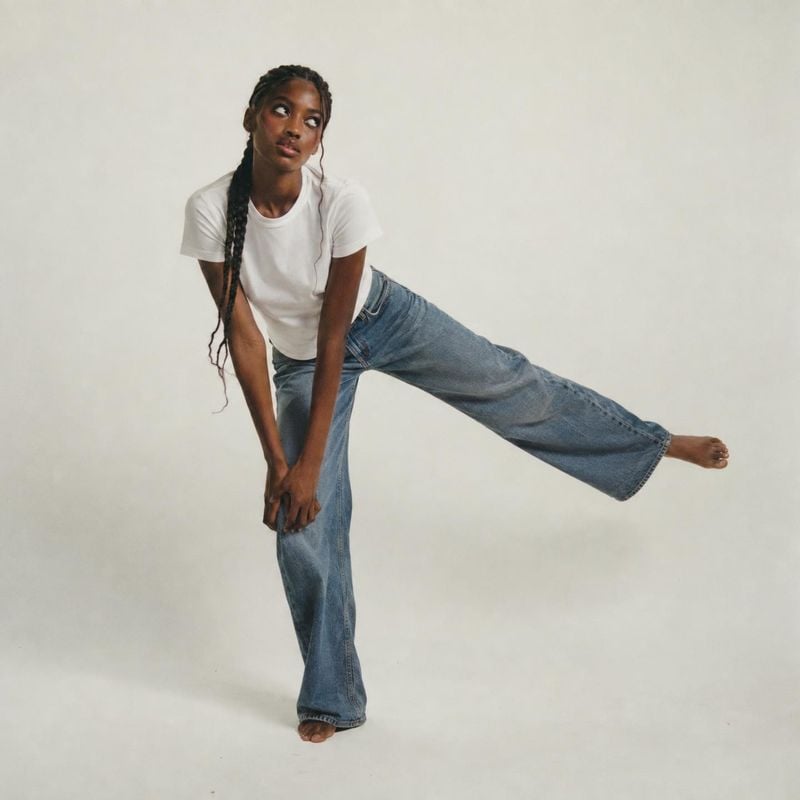 blue jeans model try-on product photo: A model in a white T-shirt and loose blue jeans balances with one leg extended against a softly lit neutral studio backd…