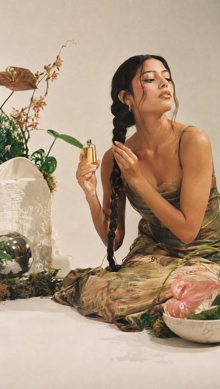 hair oil model try-on product photo: A woman in a flowing botanical-print dress holds a small golden hair oil bottle while tending to her long braid, posed amo…