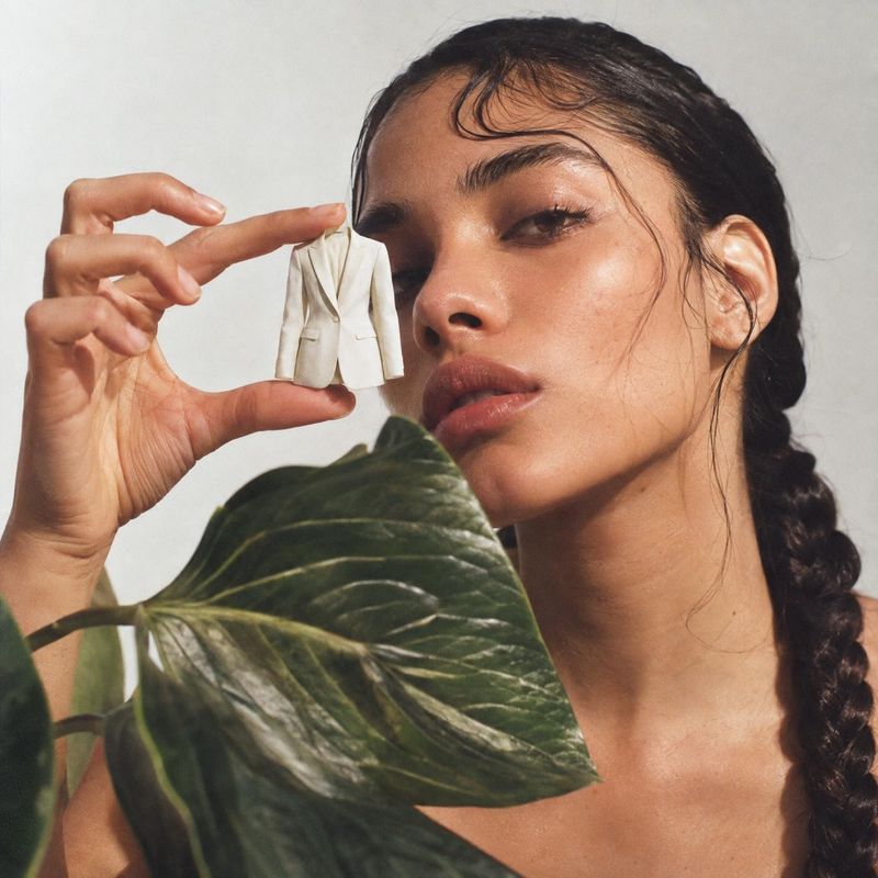 miniature blazer product in hand product photo: A close-up fashion portrait shows a woman holding a tiny ivory blazer beside her face, with a large green leaf…