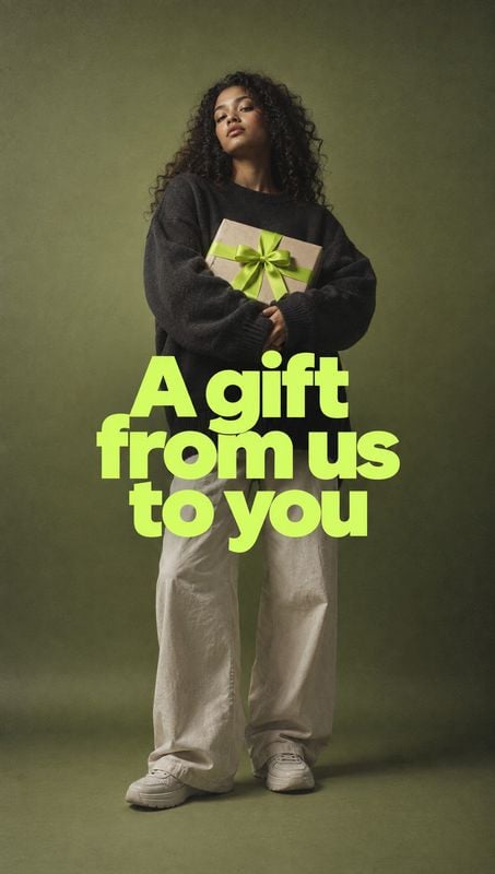 gift box model try-on product photo: A woman in a charcoal sweater, loose light trousers and sneakers holds a ribbon-wrapped gift against an olive studio backd…