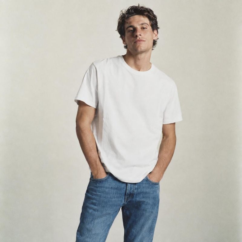 white T-shirt model try-on product photo: A casually posed male model wears a plain white T-shirt and blue jeans against a softly lit neutral studio backdrop,…