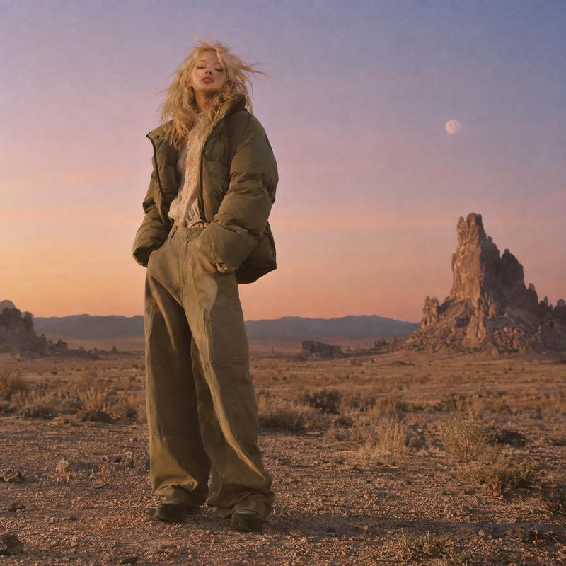 puffer jacket and trousers action & motion product photo: A model wears a dark padded jacket and loose trousers in a windswept desert landscape, lit by warm su…