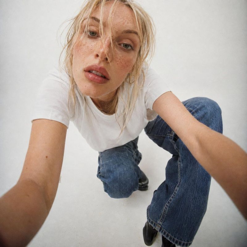 casual outfit model try-on product photo: A blonde model in a white T-shirt, blue jeans, and black shoes leans toward the camera against a bright white studio…